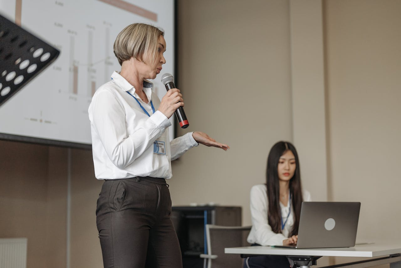 The Art of Drawing Readers In: Your attractive post title goes here Female speaker presenting at a business conference with an audience member working on a laptop.
